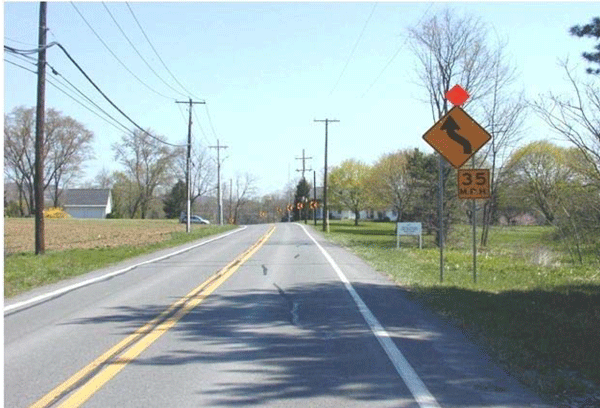 Photograph of a two-lane roadway with a horizontal curve. A horizontal curve warning sign is supplemented with a speed advisory plaque on the bottom and an orange flag on the top. The signs are installed on the right side of the roadway prior to the curve. Chevrons are also installed along the outside of the curve.