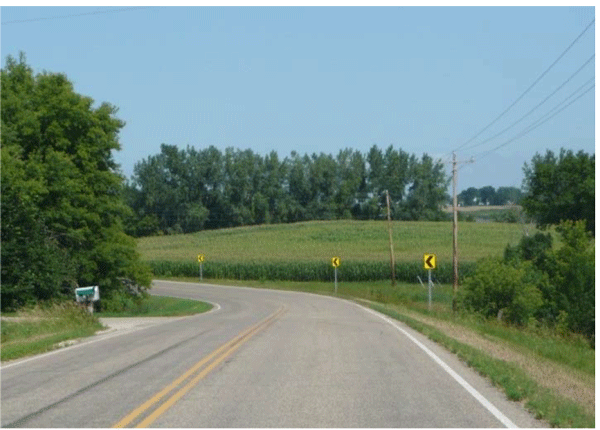 Photograph of a two-lane horizontal curve. A side road intersects the road from outside of the curve. When approaching the curve, it is difficult to see the side road.