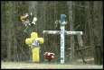 This image shows a roadside memorial marking a fatal accident involving a teenage boy. The memorial includes two crosses, a wreath, flowers, and several personal mementos.