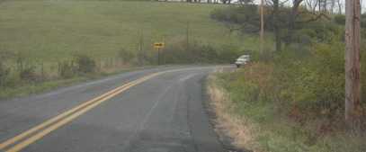Photo. Photo is a one direction large arrow sign placed on the left side of a road on the approach to a horizontal right curve.  A car can be seen rounding the curve.