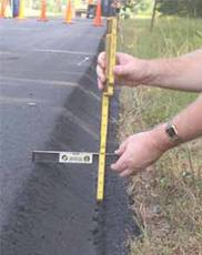 Photo.  Photo shows a set of hands at a roadway edge holing a ruler perpendicular to the roadway and a level that rests on the roadway surface.  The ruler is placed so that one end is at the lowest point of the roadway edge and the level is used to show the height deference between that point and the roadway.