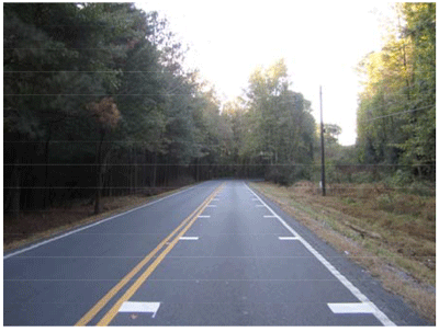 Photograph of a two-lane roadway with centerlines and edge lines in a rural area. On one side of the roadway, transverse stripes are installed between the centerline and edge line at gradually decreasing distances. The transverse stripes are perpendicular to the centerline and edge line.
