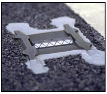 Close-up photograph of a snowplowable retroreflective raised pavement marker. The reflective device is encased in an iron casting and recessed below the pavement surface in a grooved section.