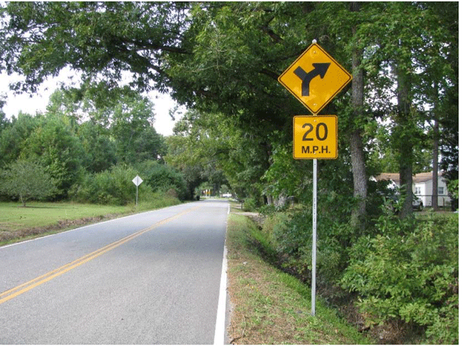 Photograph of an advisory speed plaque on one side of a two-lane roadway. The advisory speed plaque has a W1-10 arrow sign on the top of a 20 mph advisory speed sign.