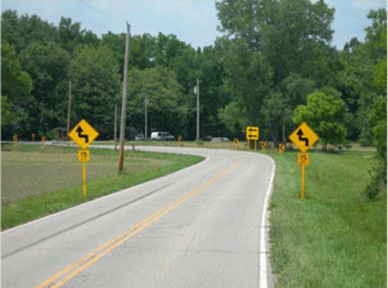 Photograph showing a curve after signage updates. This is an after period photo of figure 27. A horizontal alignment sign with an advisory speed plaque is installed on each side prior to the curve. Doubled one-direction large arrow signs are installed where there used to be only one one-direction large arrow sign.