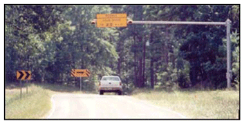 Photograph showing an overhead Texas system curve advisory speed limit sign. Chevrons and a one-direction large arrow sign are installed on the outside of the two-lane curve. An overhead curve advisory speed limit sign is installed on the inside of the curve.