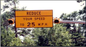Close-up photograph of the Texas system curve advisory speed limit sign, which is showing in Figure 34. A rectangular warning sign is mounted on an arm of a utility pole with the text reduce your speed to 25 mph on the sign. There are two flashing beacons. One is mounted on the right side and the other one is mounted on the left side.