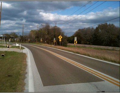Photograph showing high friction surface treatment on a horizontal curve of a two-lane roadway. Chevrons and horizontal curve warning signs are installed on the outside of the curve. The roadway has centerlines and edge lines. Pavement in the middle is darker than the surrounding pavement. The darker layer of pavement in the photograph is an application of high friction surface treatment.