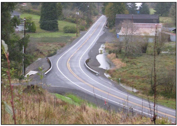 Photograph showing a road intersecting a two-lane curve. Guardrails are installed on inside and outside of the curve. A portion of the approach is paved.