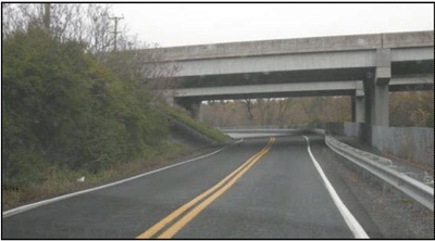 Photograph of a two-lane roadway with a guardrail on the right and an embankment on the left. A bridge crosses over the roadway. A white edge line is installed on both sides to define and delineate the edge of the roadway.