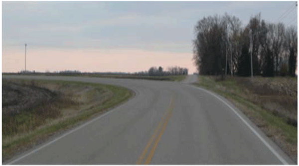 Photograph of a two-lane horizontal curve. A side road intersects the road from outside of the curve. When approaching the curve, it is difficult to see the side road.