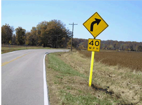 White edge lines are installed on both sides of the curve to define the edge of the roadway surface. No shoulder is present for this roadway. A horizontal curve warning sign supplemented with an advisory speed plaque is installed on the right side prior to the curve.