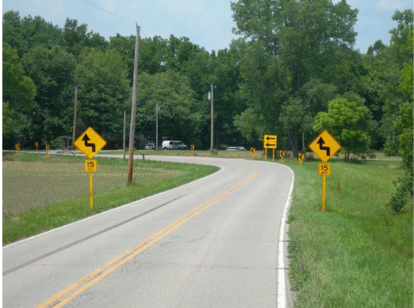 Photograph of the same curve as shown in Figure D-1 after signage updates. In the photo, one-direction large arrow sign is doubled. Chevrons are also installed on the outside of the curve. A horizontal curve waring sign supplemented with an advisory speed plaque is installed on both sides of the roadway prior to the curve.