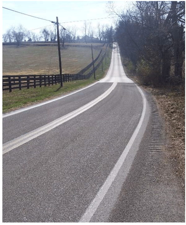 Photograph of a rural, two-lane roadway with a horizontal curve. A darker layer of pavement is present within the curve, between the centerline and edgeline, which is the application of the HFST.