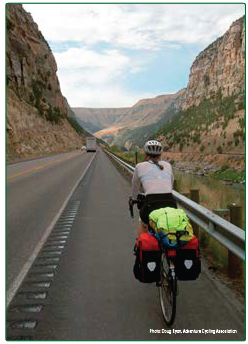 A bicyclist travelling on a wide shoulder that is edged on the left with rumble strips that contain gaps through which a cyclist may move without being forced to traverse the rumble strips themselves. Photo credit: Doug Ryan, Adventure Cycling Association.