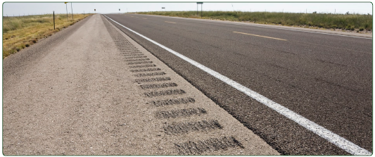 A long, straight roadway in a rural area featuring rumble strips on the shoulders.