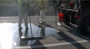 A spiggot releasing binder compound onto the roadway, where a worker uses a broom to spread it evenly over the surface.