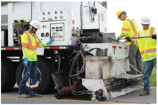 Workers monitor a truck that combines and spreads aggregate and applies and smooths binder autonomosly.