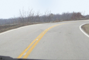 The photo shows US 60 in the segment known as the incidental section.  The roadway is a two-lane rural road with no shoulders and horizontal and vertical curves.  A guardrail is provided to protect drivers from a steep embankment on one side.