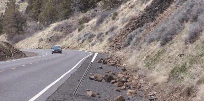 Figure 16: Photo of a car travelling along a horizontal curve toward the viewer. A mile marker sign is bent and steep terrain is on the right hand side. Fallen rock lays along the side of the road.
