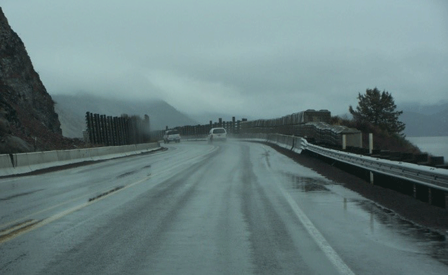 Figure 18: Photo of two vehicles travelling along a horizontal curve in rainy and cloudy conditions. The road is wet with guardrails and concrete barriers on each side. Fog and clouds reduce visibility.