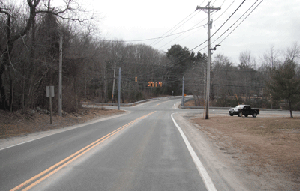 Photo of the Snake Hill Road and Chopmist Road intersection looking west, showing the horizontal curve west of the intersection (far-side). On the near-side of the intersection (east side) a utility pole and span wire pole are located in the clear zone. A power line pole and a traffic signal is seen in the distance along with a truck parked on the side of the road.