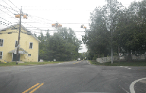 Photo of the intersection of Snake Hill Road and Sawmill Road showing flashing signal heads hanging with power lines and a yellow house in the background.
