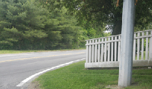Photo of a wooden fence obstructing the sight lines along the southern leg of the intersection. A pole is in the front right portion of the photo and trees and vegetation are in the background.