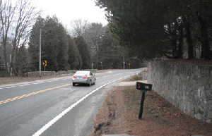 Photo showing a car driving away from the viewer with a mailbox on the right side with a retaining wall behind it.  Another retaining wall and a utility pole are on the other side of the road.