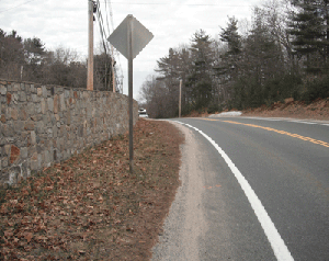 Photo of a road curving to the left. A retaining wall and crest limit sight lines along the northern edge of the curve.  The back side of a roadway sign is seen in the middle of the photo with a white painted line and double yellow centerline to the right hand side of the page.
