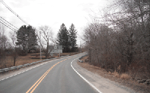 Photo of Snake Hill Road in a Westbound direction approaching a curve. A guardrail is on the left and a house is in the distance with trees along the right side. Some trees are on the left side as well.