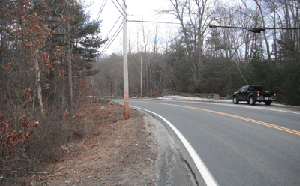 Photo of Snake Hill Road approaching a curve in the westbound direction. Trees are on the left side, a utility pole in the middle, white painted line, double yellow centerline and a truck parked on the side of the road. The pavement edge along the inside of the curve ends in a jagged drop-off within six inches of the painted edgeline.