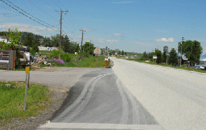 Photo of the eastbound view from the intersection of US Highway 2 and Batavia Lane. There are overhead utility lines on the left hand side. In the foreground is a small piece of blacktop pavement that has tire tracks shown where cars pull off the road.