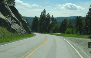 Photo of a horizontal curve near milepost 107 on US Highway 2. A rock face is on the left side, immediately adjacent to the inside curve.  A gravel pull off is located on the outside of the curve, opposite the rock face.  Just past the pull off, guardrail is located adjacent to the road.