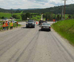 Photo of the southbound view on Springcreek Road approaching the intersection of US Highway 2. US Highway 2 is uncontrolled and Spring Creek Road is stop controlled. Skid marks are present approaching the stop sign. Construction workers in fluorescent vests are walking along the left hand side of the road. One vehicle is approaching the camera and three vehicles are faced away from the camera stopped at the stoplight. Span wire flashing beacons are located above the intersection.