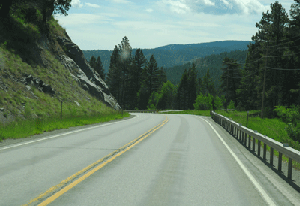 Photo of a view eastbound on US Highway 2 showing the curve near milepost 107. There is a guardrail on the right hand side of the photo and a steep hill on the left side of the road.  Mountains and trees are in the background.