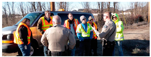 Photo of two safety patrolment and a group of people wearing retroreflective vests on a rural roadside having a discussion.
