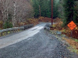 Photo of an unmarked rural roadway and a single-lane bridge over a culvert.