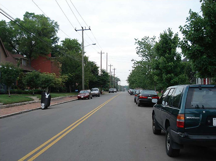 Figure 3.19.3. On-Street Parking on a Collector Street. This figure contains a picture of a two lane street divided by a double yellow line. The picture is taken from the point of view of a passenger or driver in the right hand lane. Cars are parked parallel to the curbs in both lanes. Houses are visible on both sides and there are numerous trees.