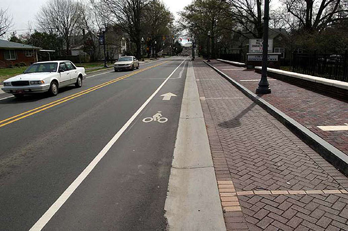 Figure 3.19.4. On-Street Parking with Adjacent Bicycle Lane. This figure contains a photograph of a two lane street divided by a double yellow line. The picture is taken from the point of view of a car. A white car and a light colored car are driving in the left hand lane. The right lane is free of traffic. To the right of the right lane, there is a bicycle lane separated from the car lane by a solid white line. A bicycle icon and arrow indicate direction of travel and the purpose of the lane. To the right of that, there are parallel parking spaces with marked dividers. The spaces are done in brick. Further to the right is a sidewalk, also done in brick, and a lamp post. A low wall separates the sidewalk from a residential area. Trees are visible in the distance.