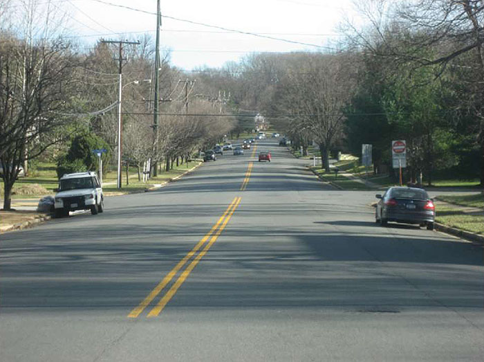 Figure 3.20.2. Example Cross-Section Prior to Road Diet. This figure contains a photograph of a two lane road separated by a double yellow line. The lanes are wide and there are cars parked on each side. Trees and power poles line each side of the street.