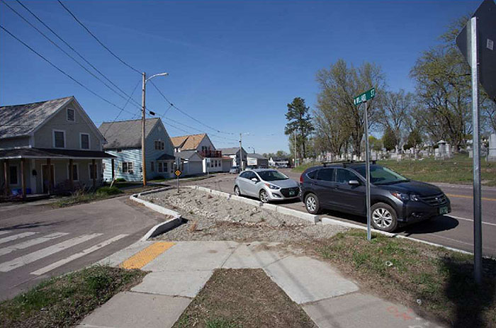 Figure 3.22.1. Full Closure at Local Street Intersection with Collector. This figure contains a photograph of a former intersection which has been closed off by a set of concrete curbs. A channel has been left for drainage and the passage of bicycles. The leg which has been closed off, running top right to bottom left in the picture, is flanked by a row of houses. The leg which runs from mid-left to bottom right of the picture has parallel parking spots, filled by two compact cars. Sidewalks can be seen near the bottom of the picture and they both lead to a crosswalk which crosses the closed off street from right to left.