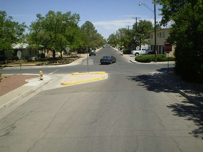 Figure 3.23.2. Half Closure Blocking Entry to Side Street. This figure contains a photograph of an undivided two lane street running top to bottom of the photo with half closure blocking entry to a side street. A separated curb extension cuts the left lane off from any oncoming traffic. A gutter between the curb and extension allows for drainage and the passage of bicycle traffic. A yellow fire hydrant is visible on the curb near the extension. A car can be seen turning from the left leg of the intersection onto the top leg. Trees and houses line the far leg. There are stop signs at each corner.