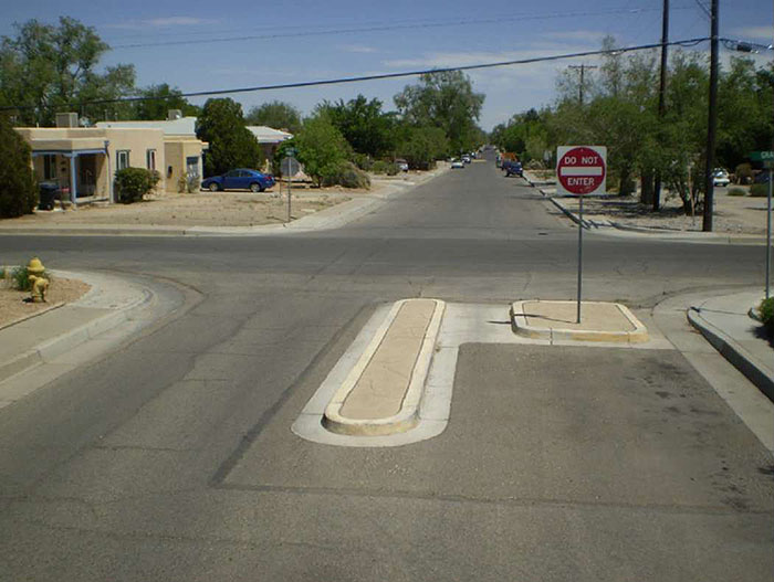 Figure 3.23.3. Half Closure Blocking Exit from Side Street. This figure contains a photograph of an intersection with half closure blocking exit from side street. The lower leg of the intersection has a square curb extension in the right lane and a small median island. A Do Not Enter sign is mounted on a signpost on the curb extension. This configuration blocks any exit from this side street. There is no traffic visible. Some street parking can be seen in the distance on the top leg.