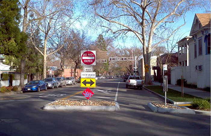 Figure 3.23.4. Half Closure Blocking Entry to Side Street with Bicycle Cut-Through. This figure contains a photograph of one leg of an intersection. The left lane uses a curb extension and a median island to stop any traffic from turning onto it. The extension and island are filled with stone. Several signs, including "Do Not Enter", "Except Bikes", and a double headed arrow pointing left to right are mounted on a single signpost on the island. The separation between extension and median allows for bike traffic. A truck is parallel parked on the right hand side. A railroad crossing is visible in the distance. Cars are also parallel parked on the left lane. Houses and trees line both sides of the street.