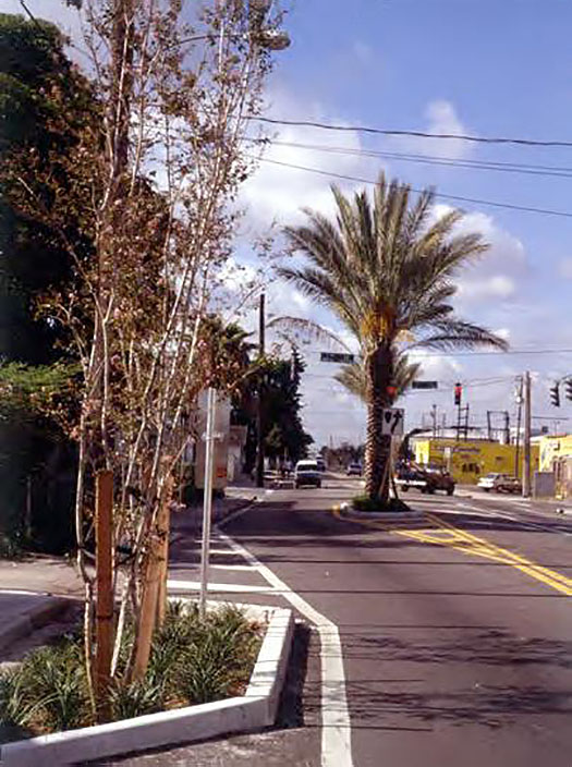 Figure 3.4.4. Lateral Shift Downstream of Signalized Intersection. This figure contains a photograph of a two lane street, showing an intersection with a traffic signal and traffic in the distance. The left lane of traffic shifts around a median containing a palm tree.