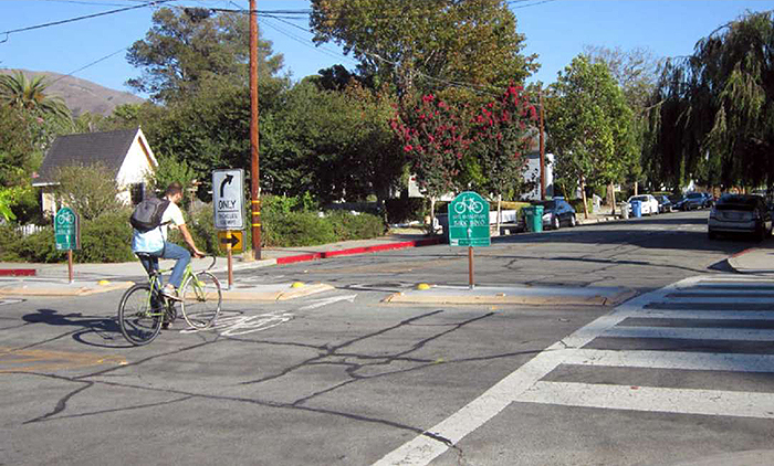 Figure 3.24.3. Median Barrier with Bicycle Boulevard Cut-Through. This figure contains a photograph of a median barrier running left to right. The street crossing this barrier allows for bicycles to travel through using cut throughs in the barrier. A cyclist can be seen using one such cut through travelling from the lower leg to the upper leg. A green bicycle sign is on one portion of the median. A "Right Turn Only" sign and curving right arrow sign are mounted on another section, showing cars that they are not able to cross over. Numerous trees and houses can be seen on the corner opposite where the picture was taken from.