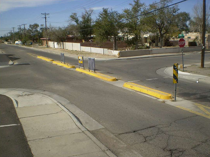 Figure 3.24.4. Narrow Median Barrier with Cut-Throughs. This figure contains a photograph focusing on one street running from bottom right to top left of the picture. A narrow concrete median barrier blocks motor vehicle traffic from a cross street. Numerous cut-throughs allow for bicycles to pass. Yellow markers and turn only signs show motor vehicle traffic how to proceed. There are trees in a lot opposite the corner where the picture was taken from.