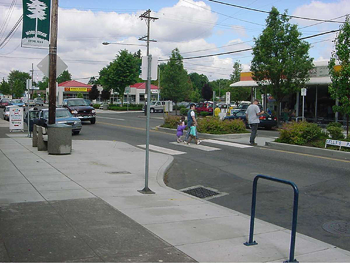 Figure 6.3. Crosswalk with Bulbout and Median Refuge. This figure contains a photograph of a commercial area of a town with a two-lane road, narrowed at a crosswalk with bulbout and median refuge with landscaping. Pedestrians are seen crossing the street, with cars waiting.