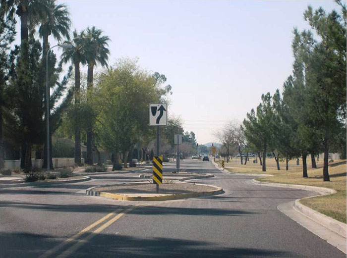 Figure 3.5.2. Chicane with Median. This figure contains a photograph of a chicane achieved using a series of circular medians. The streets are lined with palm trees and each median holds a sign post with a square sign indicating right of way via a curved arrow and underneath that a rectangular sign with yellow and black diagonal stripes.